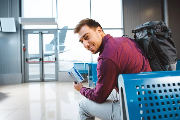 Enfoque selectivo de sonriente hombre con pasaporte con billete de avión y sentado cerca de la puerta en el aeropuerto - foto de stock