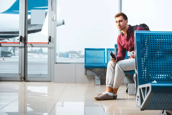 Bel homme tenant smartphone et passeport tout en étant assis dans le salon de départ — Photo de stock