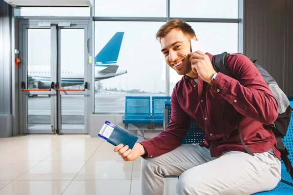 Улыбающийся мужчина разговаривает на смартфоне, сидя в аэропорту с рюкзаком — стоковое фото