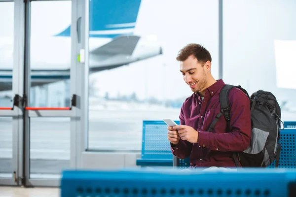 Homem alegre usando smartphone e sorrindo enquanto espera na sala de embarque — Stock Photo