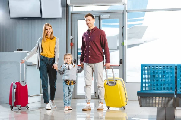 Happy mom and dad holding hands with daughter and standing with suitcases near gate in airport — Stock Photo