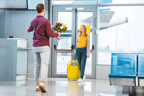Вид ззаду хлопця з квітами зустріч щаслива дівчина з валізою в аеропорту — стокове фото