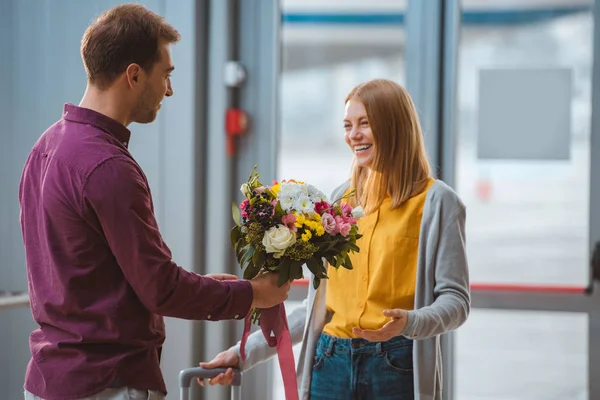 Красивый бойфренд с цветами во время встречи со счастливой подругой в аэропорту — стоковое фото