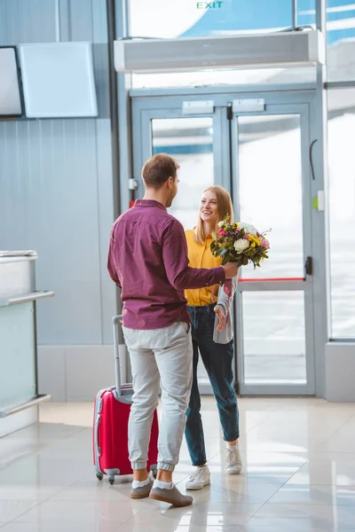 Бойфренд держит цветы и встречает счастливую девушку с чемоданом в аэропорту — стоковое фото