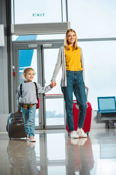 Attractive mother holding hands with cute daughter while walking with suitcases in airport — Stock Photo