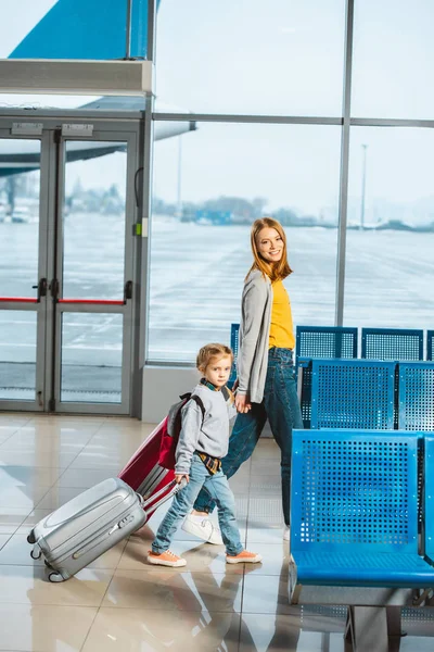 Atractiva madre cogida de la mano con su hija y caminando con equipaje en el aeropuerto - foto de stock