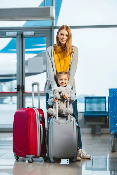 Alegre madre abrazando linda hija sosteniendo osito de peluche cerca de equipaje en aeropuerto - foto de stock