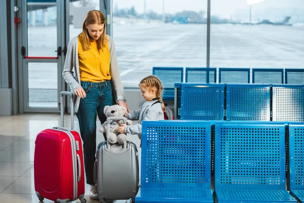 Mère debout avec fille mignonne et regardant ours en peluche sur les bagages à l'aéroport — Photo de stock