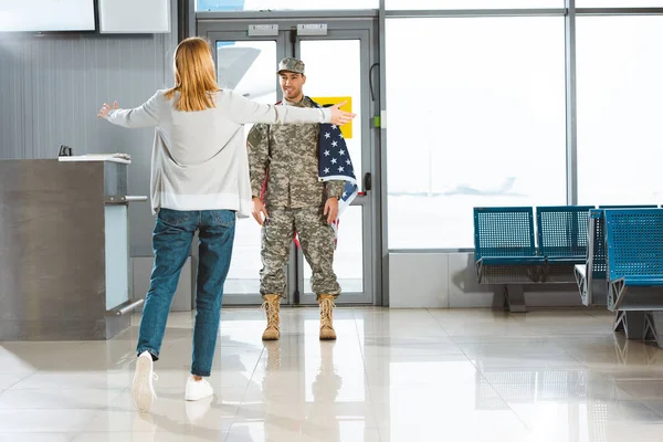 Novia feliz con los brazos abiertos reunión novio en uniforme militar con bandera americana en el aeropuerto - foto de stock
