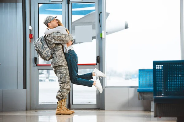 Feliz novio en uniforme militar celebración en brazos novia en aeropuerto - foto de stock