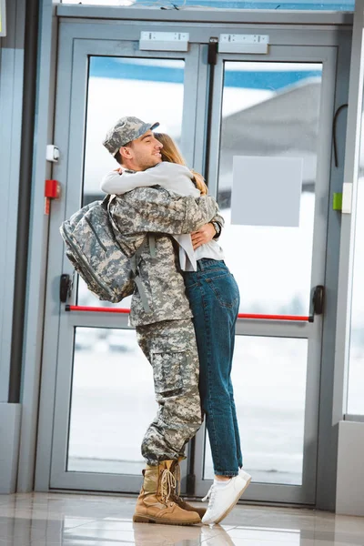 Happy girlfriend hugging cheerful boyfriend in military uniform in airport — Stock Photo