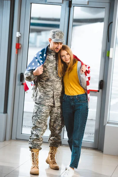 Happy veteran in military uniform standing with girlfriend and holding american flag in airport — Stock Photo