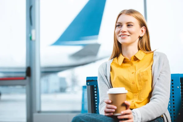 Hermosa mujer sosteniendo taza de papel mientras está sentado en el aeropuerto - foto de stock