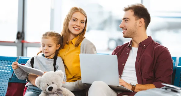 Cute daughter holding digital tablet near mother and sitting near father with laptop in airport — Stock Photo