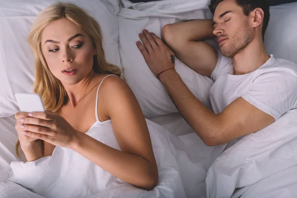 Top view of suspicious young woman using smartphone and looking at boyfriend sleeping in bed — Stock Photo