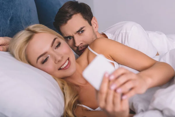 Joven sospechoso espiando a su esposa sonriente usando el teléfono inteligente en la cama, concepto de desconfianza - foto de stock