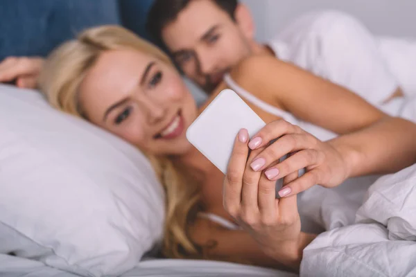Enfoque selectivo del joven que mira a su novia sonriente usando el teléfono inteligente en la cama, concepto de desconfianza - foto de stock