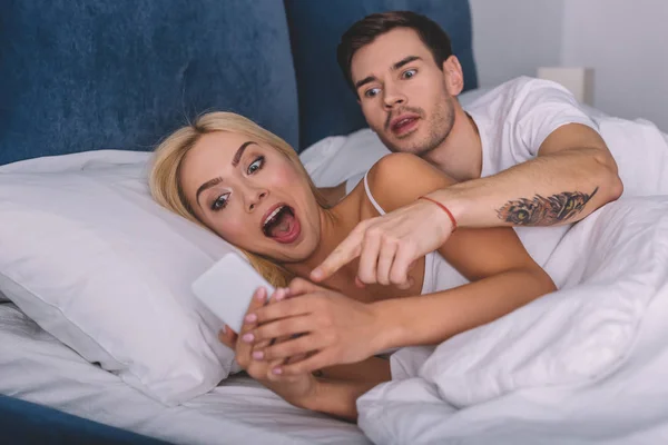 Shocked young woman holding smartphone and emotional man pointing at screen while lying together in bed — Stock Photo
