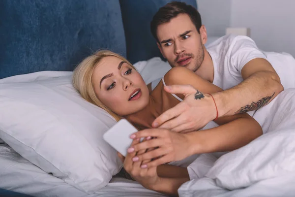 Angry man looking at suspicious girlfriend using smartphone in bed, mistrust concept — Stock Photo