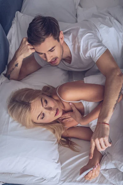 Top view of young woman using smartphone and emotional man pointing at screen in bed, secret concept — Stock Photo