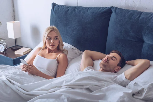 Vue grand angle de belle femme en utilisant un smartphone et en regardant dormir mari au lit — Photo de stock