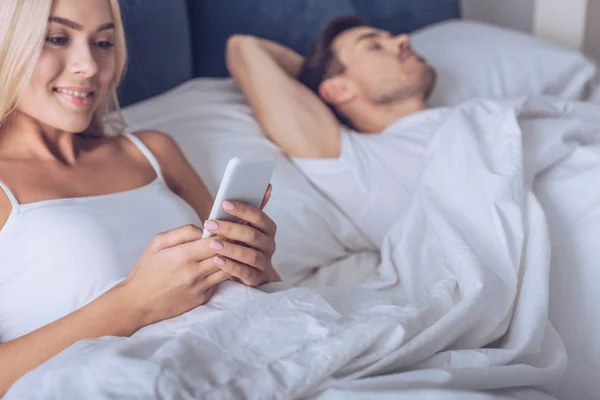 Hermosa mujer joven sonriente usando teléfono inteligente mientras está acostado en la cama con el marido dormido - foto de stock