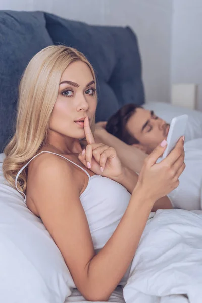 Jovem mulher gesticulando para o silêncio e olhando para a câmera enquanto namorado dormindo na cama, conceito secreto — Fotografia de Stock