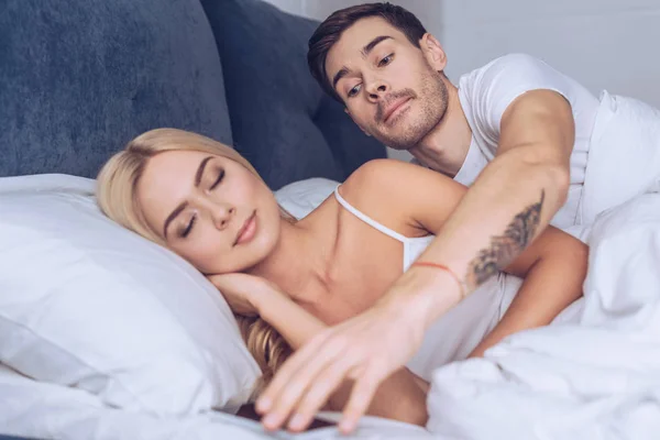 Suspicieux jeune homme atteindre pour smartphone tandis que belle petite amie dormir dans le lit, concept de méfiance — Photo de stock