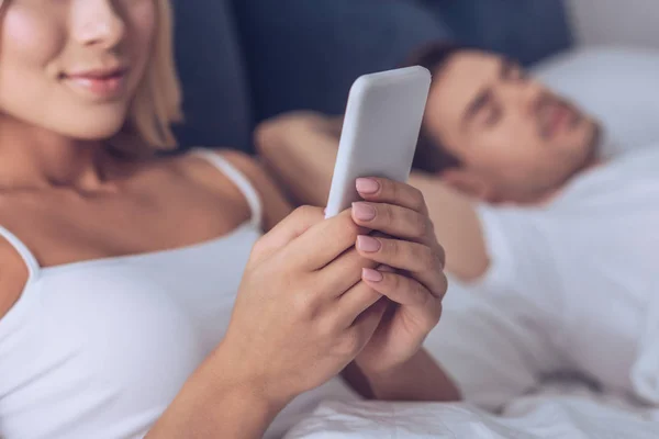 Schnappschuss einer lächelnden jungen Frau mit Smartphone, während ihr Mann im Bett schläft — Stockfoto