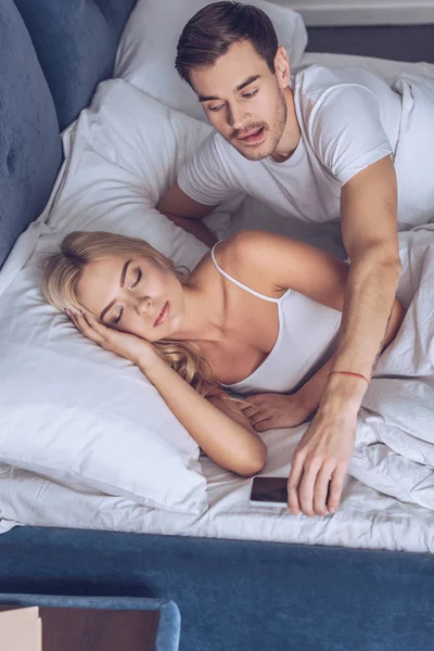 Vue grand angle du jeune homme jaloux atteindre pour smartphone tandis que la femme dort au lit, concept de méfiance — Photo de stock
