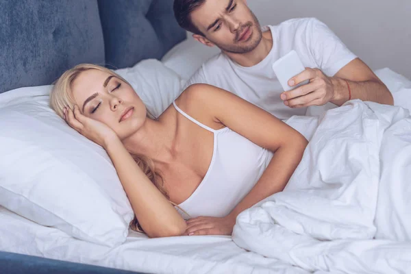 Підозрілий молодий чоловік використовує смартфон, коли дівчина спить у ліжку, секретна концепція — стокове фото