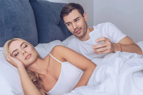 Sonriente joven sosteniendo teléfono inteligente y mirando a dormir novia en la cama, concepto secreto - foto de stock