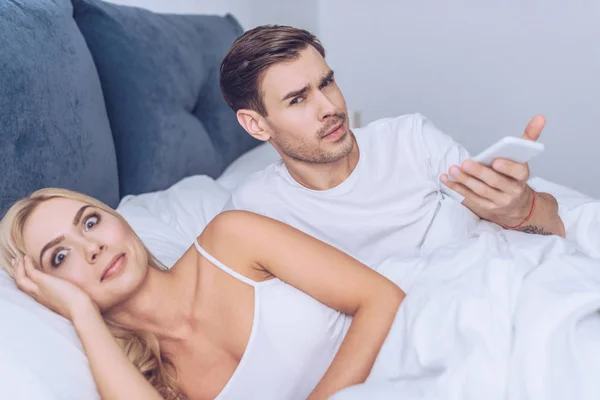 Jealous young man holding smartphone and looking at camera while lying with shocked wife in bed — Stock Photo