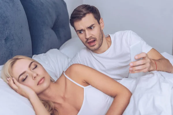 Joven enojado sosteniendo el teléfono inteligente y mirando a la novia dormida en la cama - foto de stock