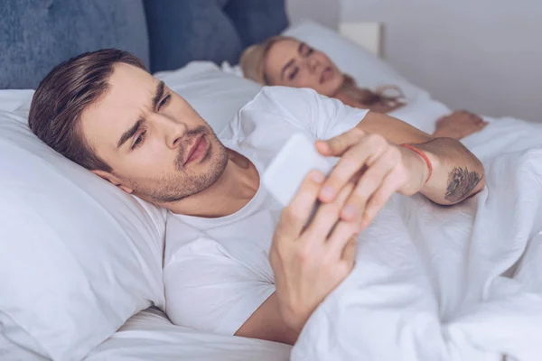 Suspicious young man using smartphone while lying with sleeping girlfriend in bed — Stock Photo