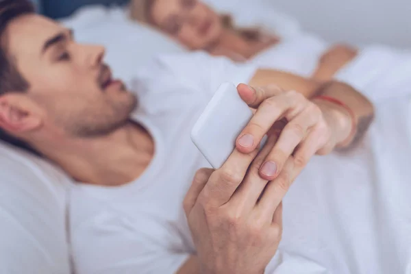Close-up view of young man using smartphone while lying with sleeping wife in bed — Stock Photo