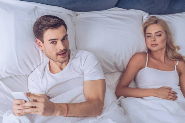 Vista de ángulo alto de un hombre joven usando un teléfono inteligente y mirando a la cámara mientras está acostado con su esposa dormida en la cama - foto de stock