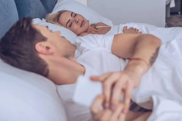 Suspicious young man using smartphone and looking at sleeping girlfriend in bed, secret concept — Stock Photo