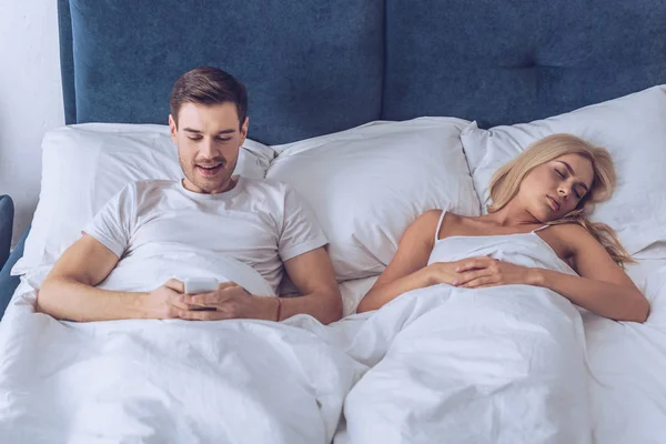 Lächelnder junger Mann, der im Bett liegt und Smartphone benutzt, während junge Frau schläft, geheimes Konzept — Stockfoto