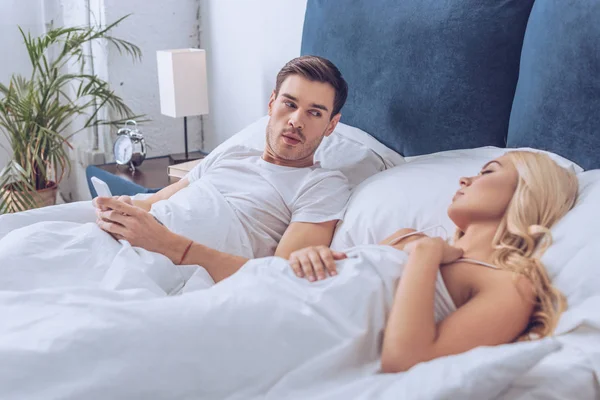 Suspicious man lying in bed with smartphone and looking at sleeping girlfriend, mistrust concept — Stock Photo