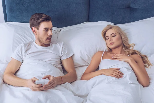 Подозрительный мужчина лежит в постели со смартфоном и смотрит на спящую жену, концепция недоверия — стоковое фото