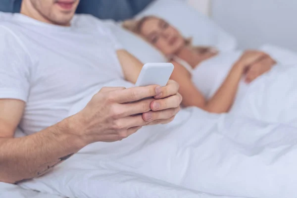 Recortado disparo de hombre joven usando teléfono inteligente mientras está acostado con la esposa dormida en la cama - foto de stock