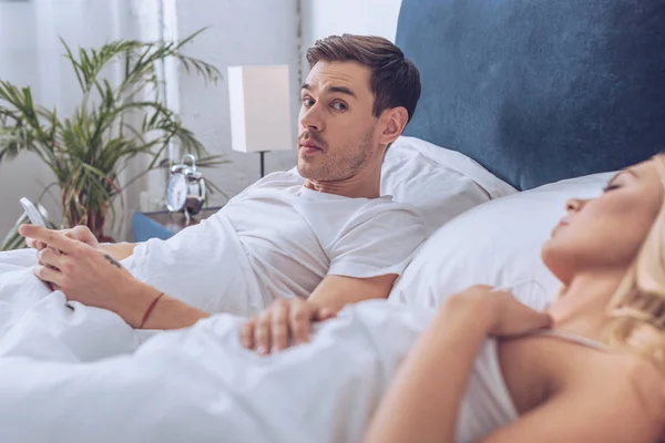 Man using smartphone and looking at camera while lying with sleeping wife in bed — Stock Photo