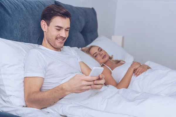 Smiling young man using smartphone while wife sleeping in bed — Stock Photo