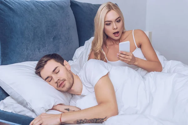 Шокована молода жінка використовує смартфон під час сну чоловіка в ліжку, недовіра концепції — стокове фото