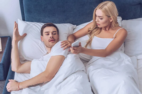 High angle view of shocked young woman showing smartphone to confused boyfriend lying in bed — Stock Photo