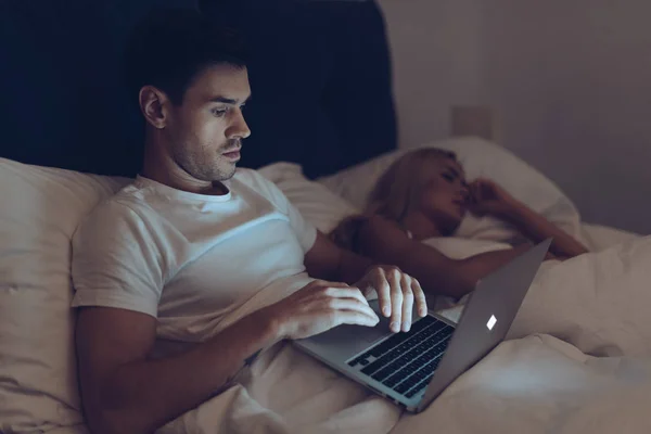 Joven sospechoso que usa el ordenador portátil mientras su novia duerme en la cama por la noche - foto de stock