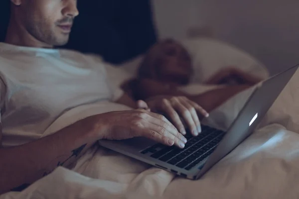 Tiro recortado de jovem suspeito usando laptop enquanto esposa dormindo na cama à noite — Fotografia de Stock