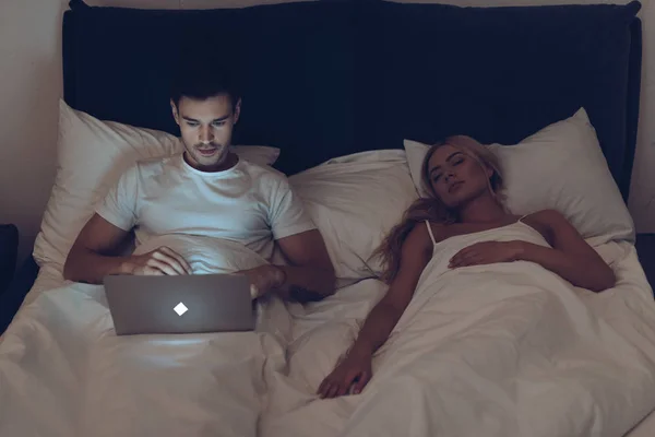 Vista de ángulo alto de hombre joven que usa el ordenador portátil mientras la esposa duerme en la cama por la noche - foto de stock