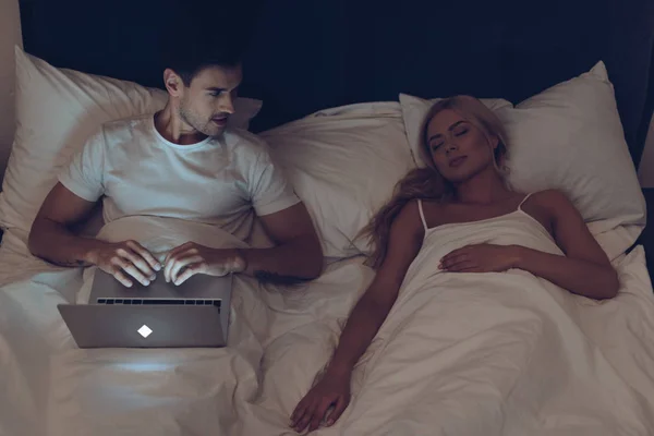 Vista de ángulo alto de hombre joven usando el ordenador portátil y mirando a la esposa dormida en la cama por la noche - foto de stock